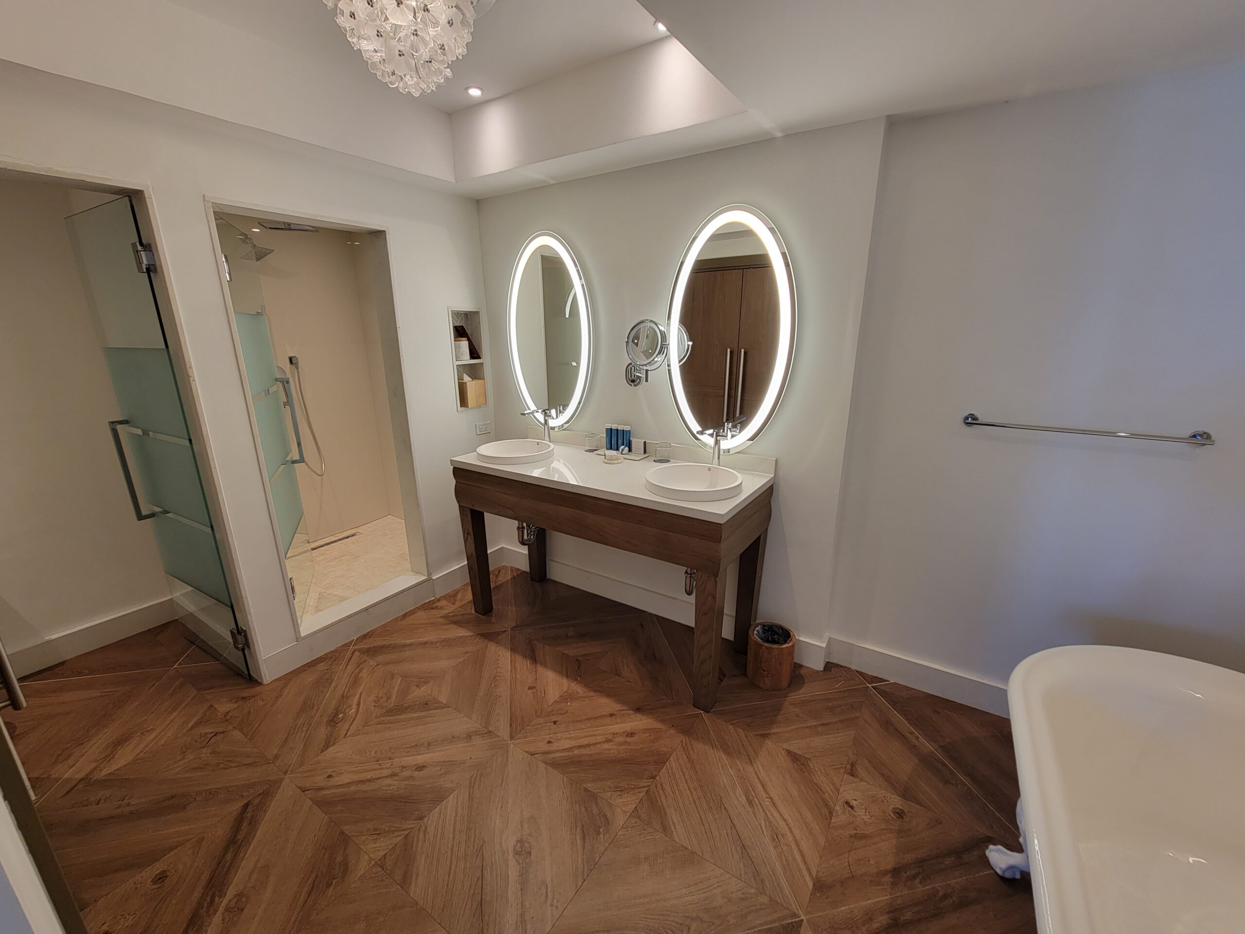 Bathroom in the Mammee Bay Beachfront One Bedroom Butler Suite (B1B) at Sandals Dunn's River, with a double vanity, twin lit oval mirrors, walk-in shower, and soaking tub