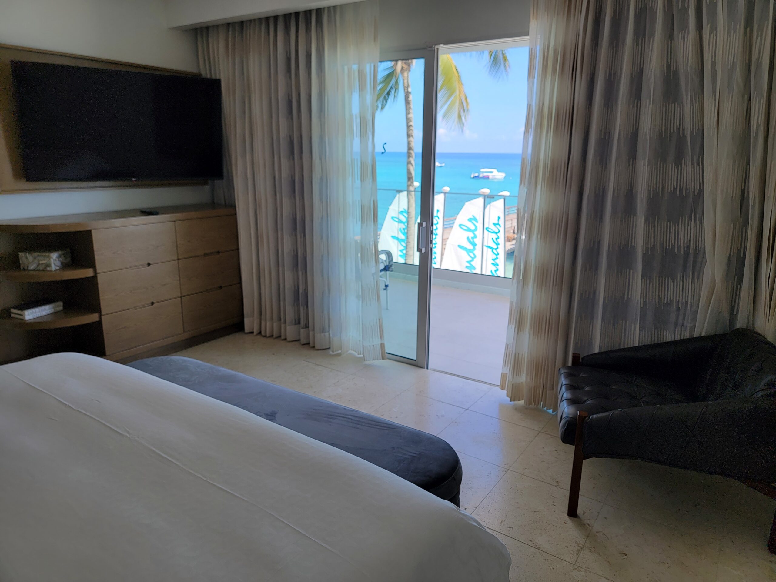 Bedroom in the Mammee Bay Beachfront One Bedroom Butler Suite (B1B) at Sandals Dunn's River, with a king bed, wall-mounted TV, and a balcony door framing a direct ocean view