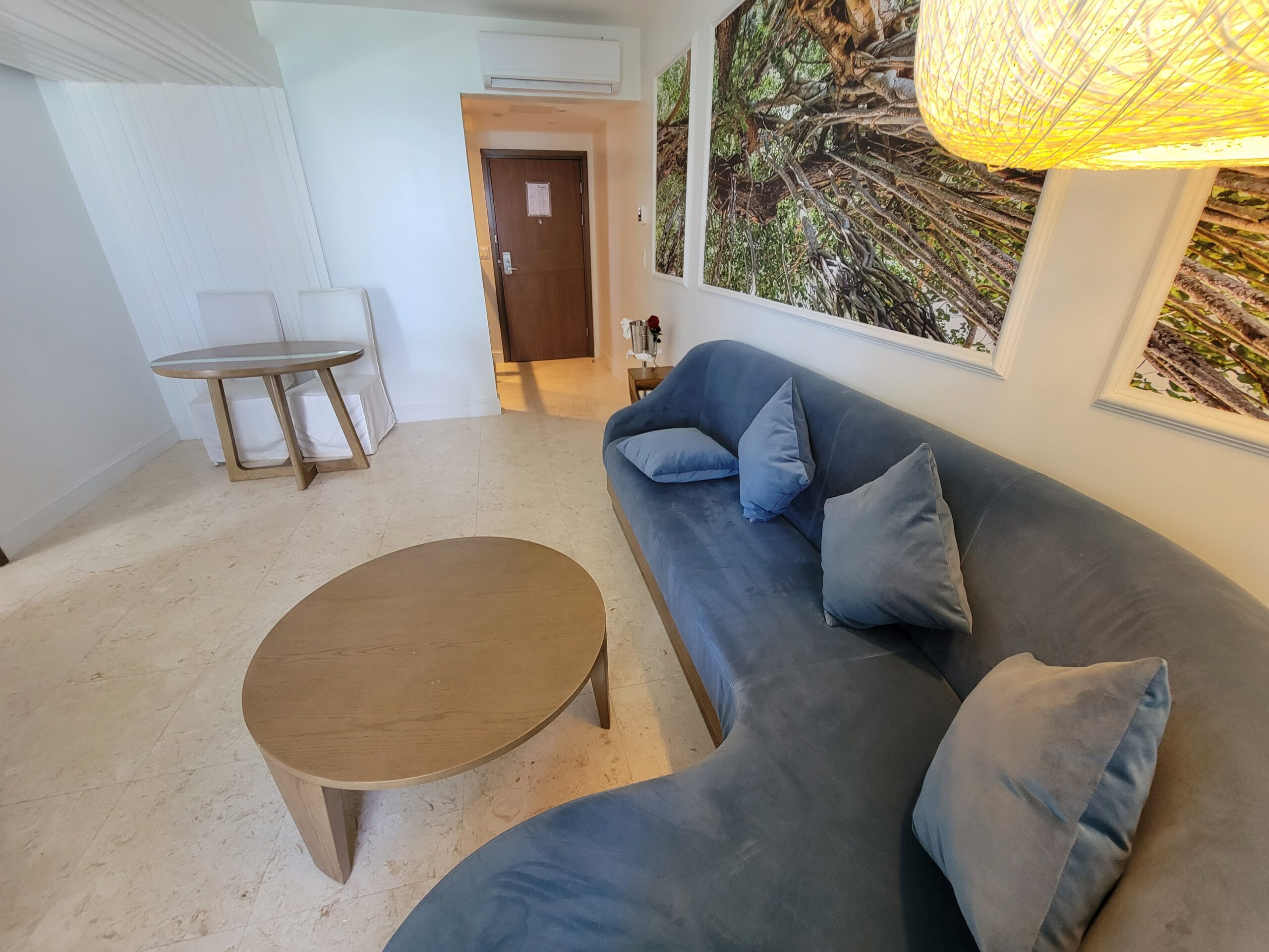 Sitting area in the Mammee Bay Beachfront One Bedroom Butler Suite (B1B) at Sandals Dunn's River, with a curved blue sofa, round coffee table, and banyan-tree wall art