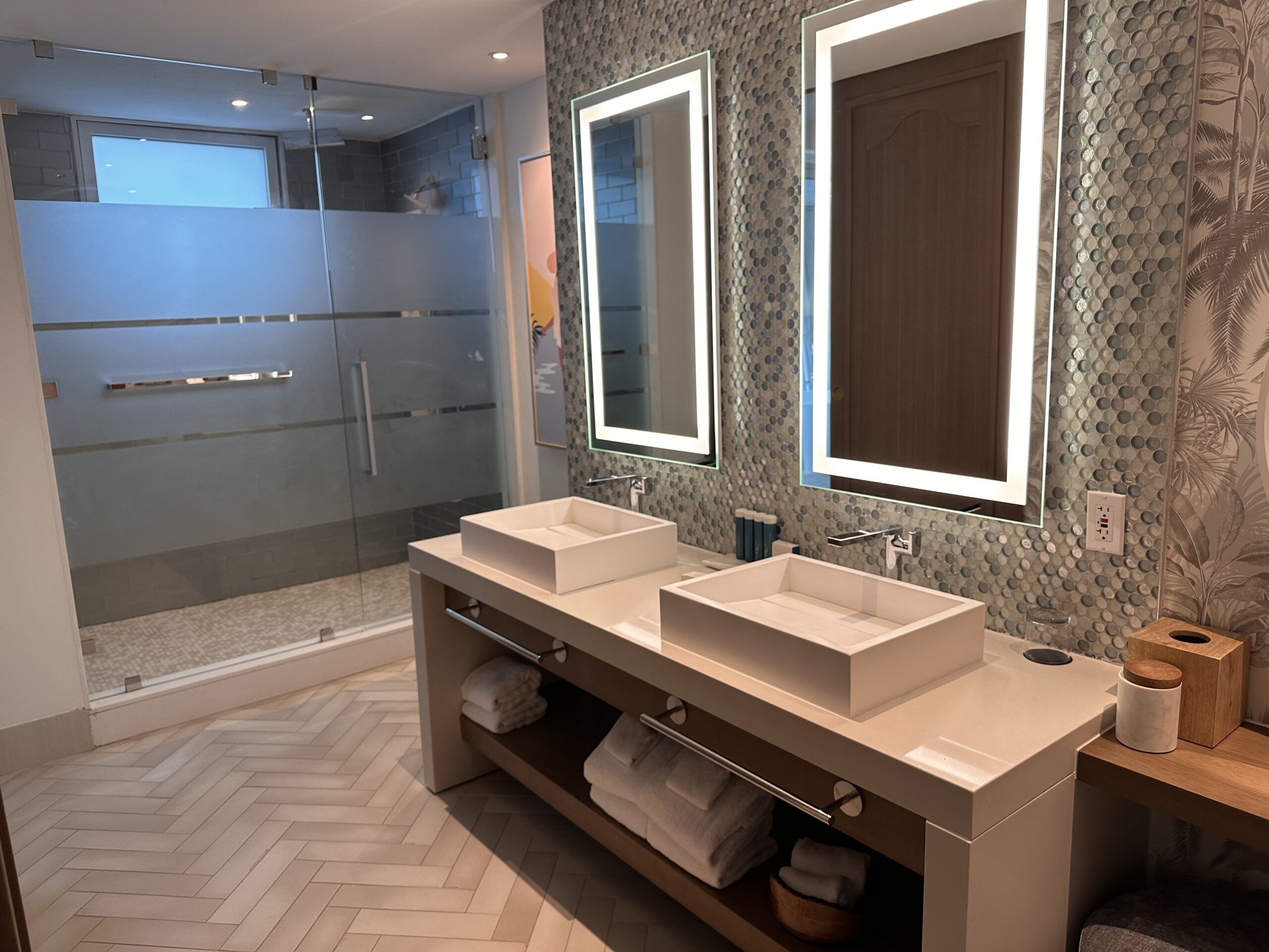 Bathroom in the Travertine Beachfront Club Level Room at Sandals Dunn's River with a double vessel sink vanity, mosaic tile backsplash, and glass-enclosed shower.
