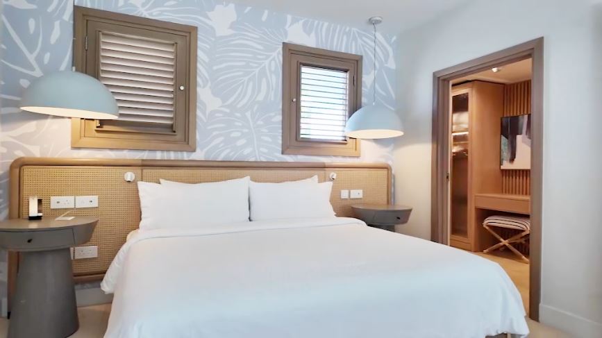 Bedroom and living area in the Butler Villa Suite with Patio Tranquility Soaking Tub (VG) at Sandals Saint Vincent, with a king bed, cane headboard, monstera-print wallpaper, and a view through to the closet