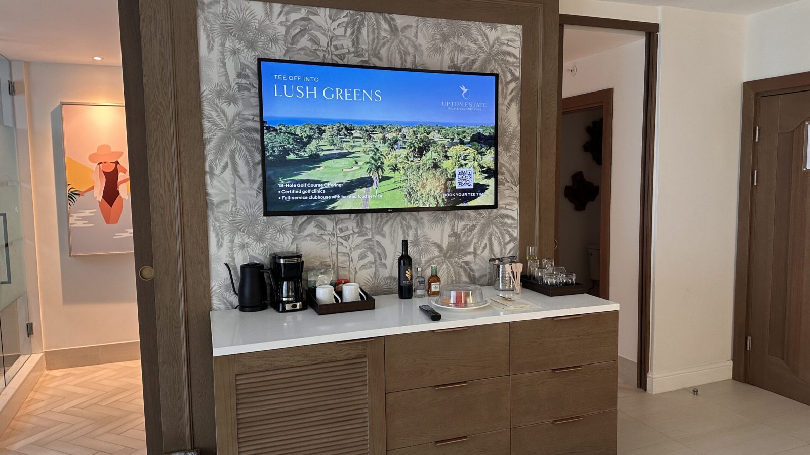 Entertainment console in the Travertine Beachfront Club Level Room at Sandals Dunn's River with a wall-mounted TV, coffee maker, and mini bar setup.
